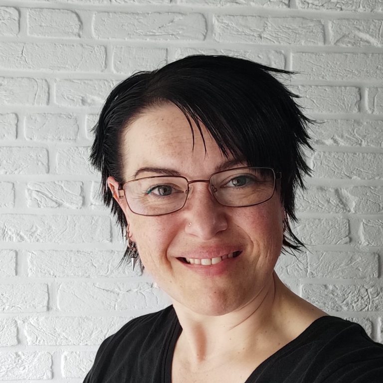 Porträt einer Frau mit kurzen, dunklen Haaren und Brille, lächelnd vor einer weißen Mauer.